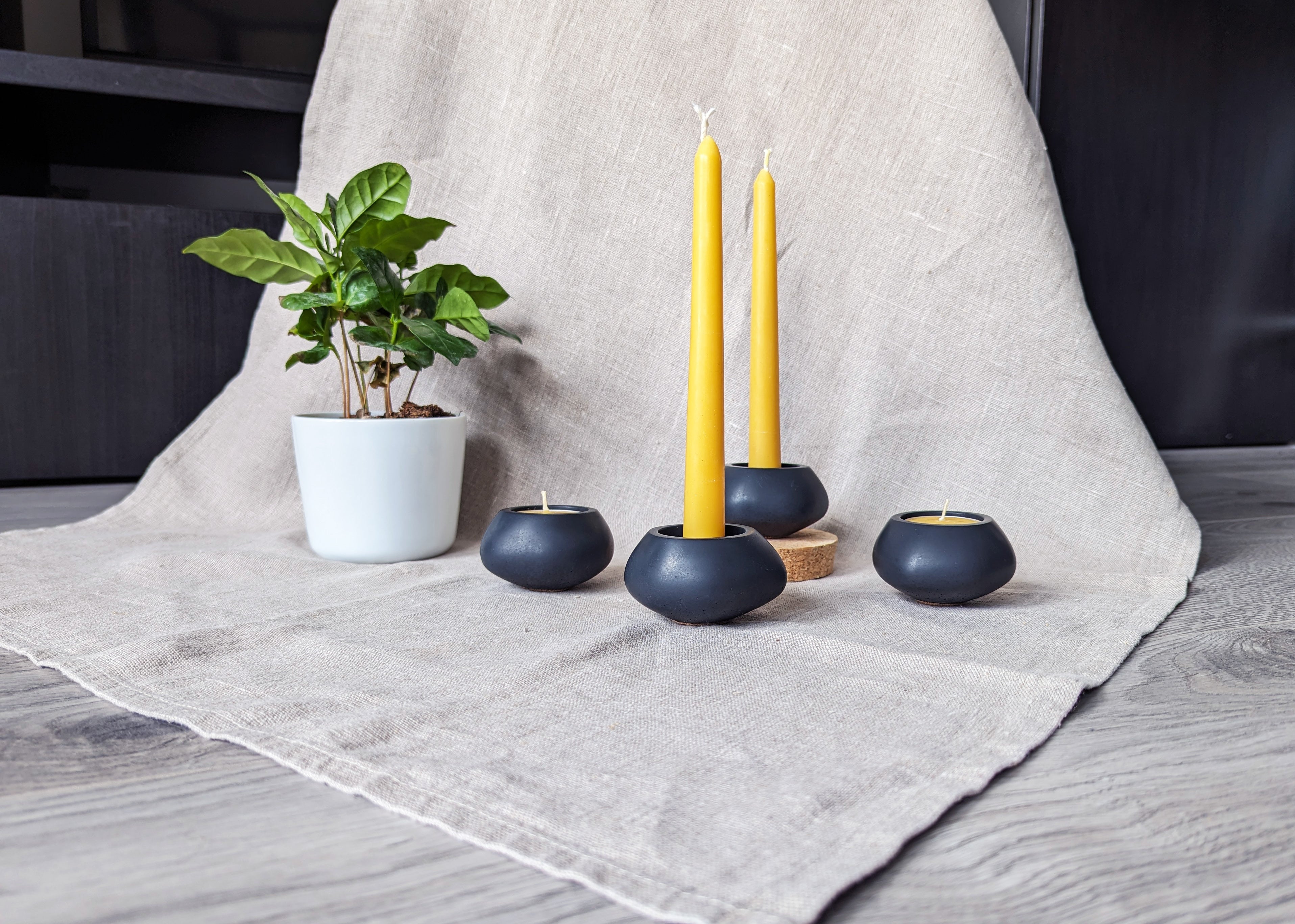 Four Votsalo candle holders holding 2 tealights and 2 dinner candles made out of beeswax, against a natural linen fabric, next to a small plant