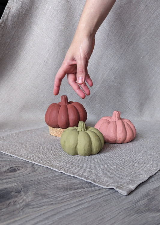 3 concrete pumpkins in 3 sizes and colours (cinnamon rose, olive green and red ochre) in a background of linen fabric. A hand is shown reaching for the pumpkins for scale.