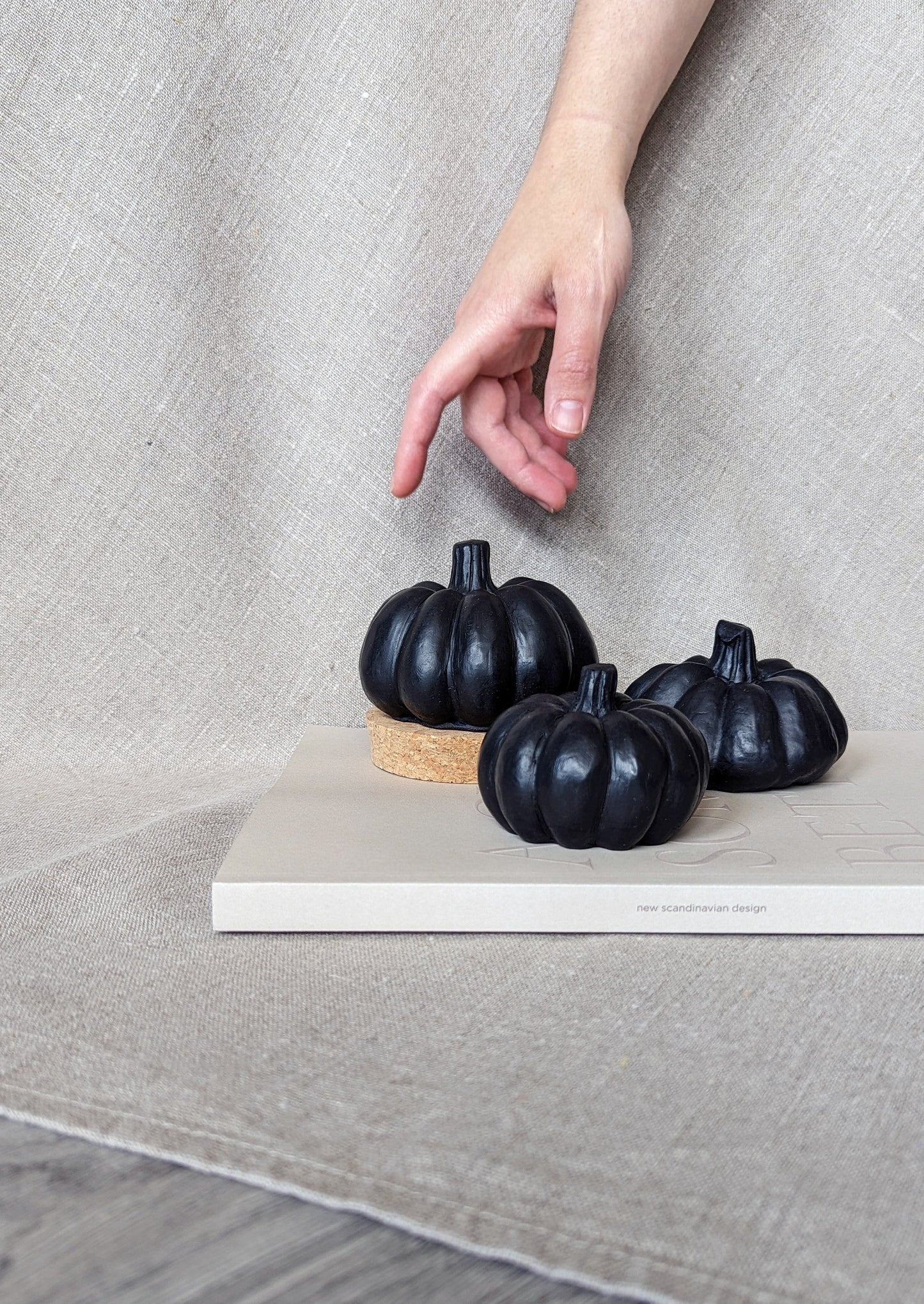 3 black concrete pumpkins in 3 sizes resting on a coffee table book, in a background of linen fabric. A hand is shown reaching for the pumpkins for scale.