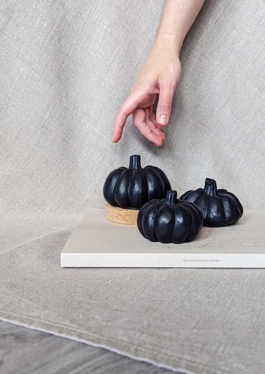 3 black concrete pumpkins in 3 sizes resting on a coffee table book, in a background of linen fabric. A hand is shown reaching for the pumpkins for scale.