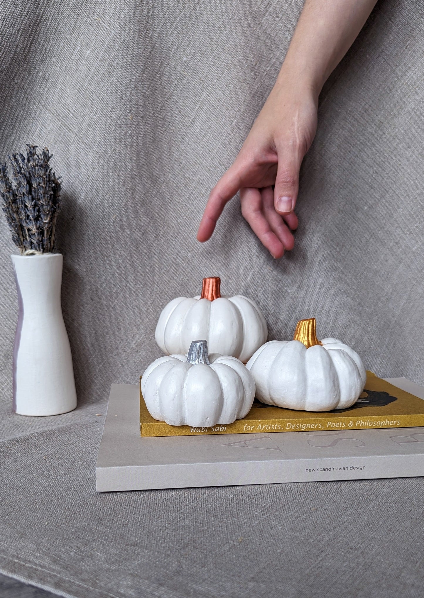 3 white concrete pumpkins in 3 sizes resting on two coffee table books, in a background of linen fabric, next to a vase with dried lavender. The pumpkins have painted tops, respectively in copper, gold and silver. A hand is shown reaching for the pumpkins for scale.