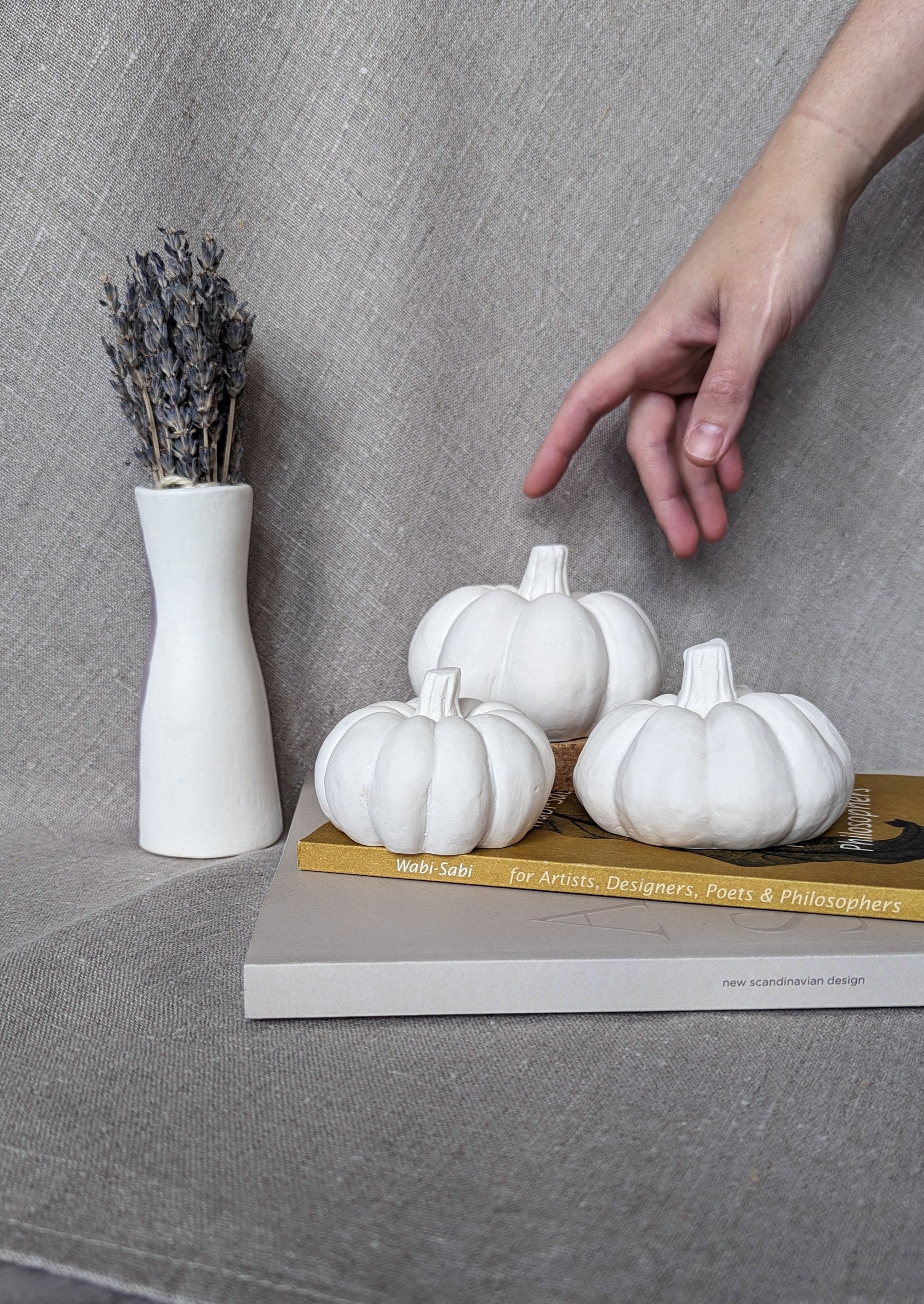 3 white concrete pumpkins in 3 sizes resting on two coffee table books, in a background of linen fabric, next to a vase with dried lavender. A hand is shown reaching for the pumpkins for scale.